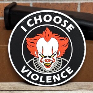 May include: A circular black patch featuring a white border and the text I CHOOSE VIOLENCE in white capital letters. The center displays a graphic illustration of a clown with bright orange hair, a white face, yellow eyes, and a ruffled collar.