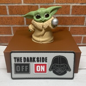 May include: A gray and black patch with the text "The Dark Side" and a toggle switch that says "Off" and "On". A black and white illustration of Darth Vader's helmet is on the right side of the patch. A green and brown toy figure of Baby Yoda is on a brown surface behind the patch.