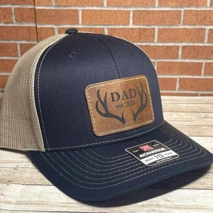 May include: A navy blue and tan trucker hat with a brown leather patch that says "DAD est. 2024" with a pair of antlers.
