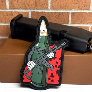 May include: A green and black bullet-shaped patch with a cartoon character holding a rifle. The character is wearing a green vest and has a white face. The patch has a red background with black bullet holes. The text "5.45" is printed on the character's vest.