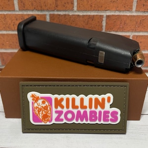 May include: A brown box with a green patch that says "KILLIN' ZOMBIES" in orange and pink letters. A black magazine with a bullet is on top of the box.