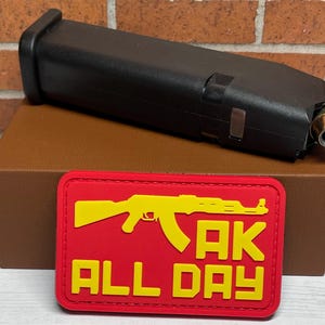 May include: A red and yellow patch with a silhouette of an AK-47 rifle and the text "AK ALL DAY".