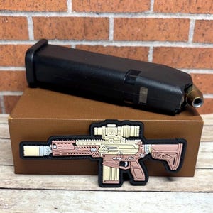May include: A black rectangular firearm magazine with a single visible cartridge resting on a brown box. Attached to the front of the box is a flat, rubberized patch featuring a detailed graphic of a rifle in shades of tan, brown, and cream.