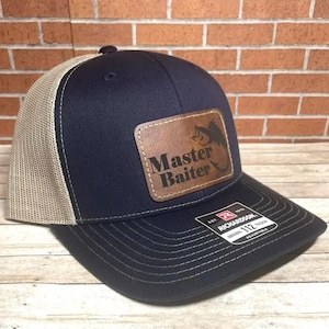 May include: A navy blue and tan trucker hat with a brown leather patch that says "Master Baiter" with a fish graphic. The hat is made by Richardson.