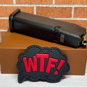 May include: A black plastic magazine with a silver metal insert and a gold-colored bullet. The magazine is sitting on a brown box with a black patch that says "WTF!" in red letters.