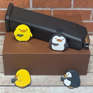 May include: Four enamel pins featuring a yellow chick and three penguins holding knives. The pins are displayed on a brown box with a black gun magazine on top.