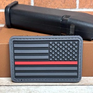 May include: A rectangular patch featuring a gray American flag design with a red stripe. The flag has gray stripes and white stars. A black object is in the background.