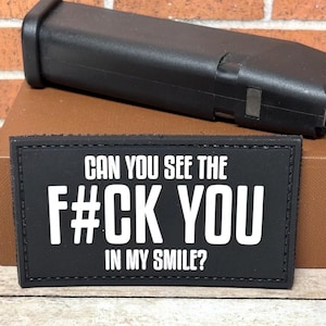 May include: Black rectangular patch with white text that reads "CAN YOU SEE THE F#CK YOU IN MY SMILE?" A black object is in the background. The patch is on a brown surface.