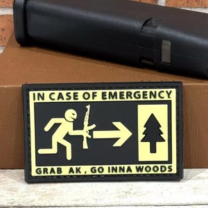 May include: Black and yellow patch with the text "IN CASE OF EMERGENCY" and an illustration of a person running with a rifle, an arrow pointing right, and a pine tree. The text below the illustration reads "GRAB AK, GO INNA WOODS".
