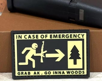 May include: Black and yellow patch with the text "IN CASE OF EMERGENCY" and an illustration of a person running with a rifle, an arrow pointing right, and a pine tree. The text below the illustration reads "GRAB AK, GO INNA WOODS".