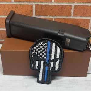 May include: A black and white skull patch with a blue stripe down the center. The skull is embroidered with a white American flag design. The patch is on a brown box.