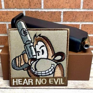 May include: A beige and brown embroidered patch with a cartoon monkey covering its ears with a gun. The text "Hear No Evil" is printed below the monkey.