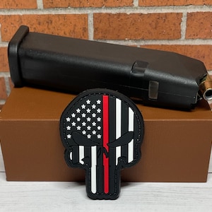May include: A black and white skull patch with a red stripe down the middle. The skull is a stylized version of the Punisher logo, with the American flag in the background. The patch is on a brown box, next to a black gun magazine.