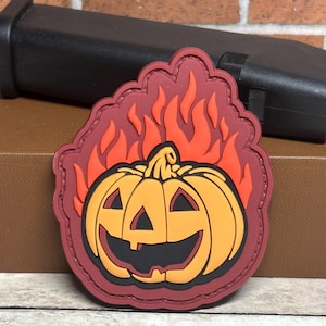 May include: A red, orange, and yellow embroidered patch of a jack-o'-lantern with flames behind it.