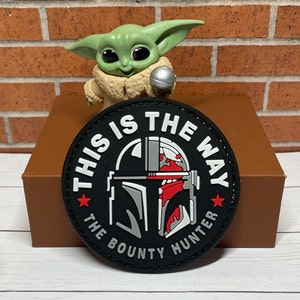 May include: A black and white patch with the text "This is the Way" and "The Bounty Hunter" around a Mandalorian helmet graphic. The patch is on a brown surface.