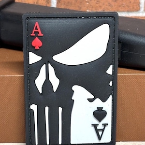 May include: A black rectangular patch featuring a white skull design, with the Ace of Spades and Ace of Hearts playing card symbols. The patch has a black border and is likely made of rubber or a similar material.