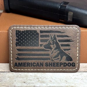 May include: A rectangular brown leather patch with white stitching around the edges. The patch features a black American flag design and a silhouette of a dog. The words "AMERICAN SHEEPDOG" are printed in black.