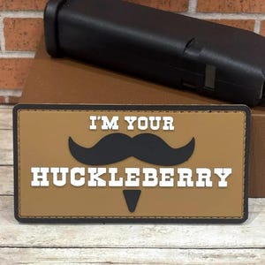 May include: A brown patch with white stitching around the edges. The patch has a black mustache and the text "I'm Your Huckleberry" in white letters.