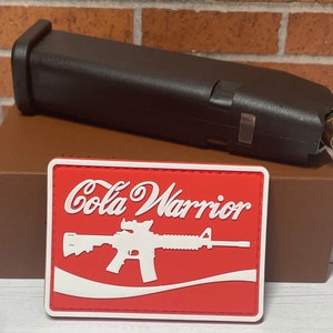 May include: A black firearm magazine with a brass-colored bullet is positioned above a red and white patch. The patch features the words "Cola Warrior" and a white graphic of a rifle.