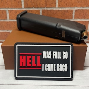May include: Black patch with white text that reads "HELL WAS FULL SO I CAME BACK" in a red and white font. The patch is on a brown box with a black magazine for a gun on top of it.