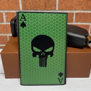 May include: Green and black patch with a skull and crossbones design. The skull is a stylized version of the Punisher logo. The patch has the letter A and a spade symbol on the top and bottom.