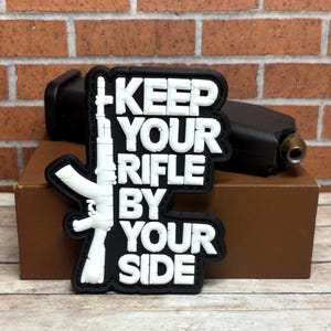 May include: A black and white patch with the text "KEEP YOUR RIFLE BY YOUR SIDE" and a white rifle graphic. The patch is on a brown box with a black object behind it, set against a brick wall.