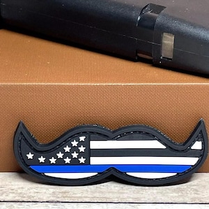 May include: Black and blue embroidered patch shaped like a mustache with an American flag and a thin blue line.