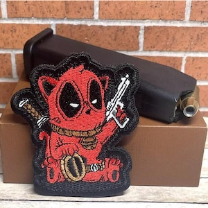 May include: A black and red embroidered patch featuring a cartoon character with a red mask and a gun. The character is wearing a gold chain and has a red and black background.