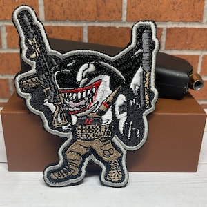 May include: A black and white embroidered patch of a shark wearing a tactical vest and holding a rifle. The shark has a menacing expression and is wearing a tactical vest with a belt and pouches. The patch is on a brown surface.