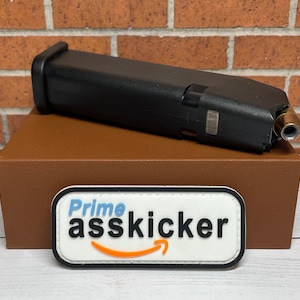 May include: A black magazine with a silver insert and a bullet in the chamber, sitting on a brown box with a white patch that says "Prime asskicker" with an orange Amazon logo.