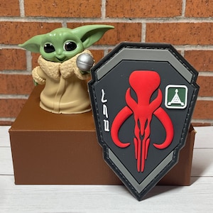 May include: A gray and red Mandalorian symbol patch with a green triangle and the text "Mando" in black. The patch is on a brown box with a green and brown toy figure in the background.