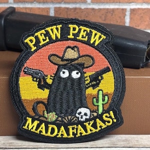 May include: A circular embroidered patch featuring a black creature wearing a brown cowboy hat, holding two black handguns in a desert landscape with a green cactus and a white skull. The text PEW PEW is at the top and MADAFAKAS! at the bottom.