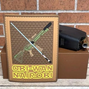 May include: A brown patch with a tan border features a green and black rifle with a blue lightsaber crossing it. The text "OBI-WAN NAIROBI!" is printed in yellow on the patch.