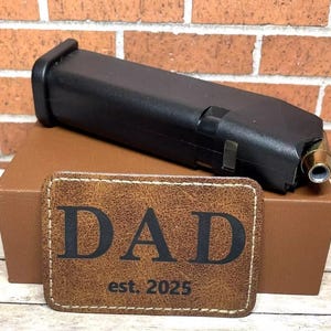 May include: A brown leather patch with the word "DAD" and the text "est. 2025" in black lettering. The patch is sitting on top of a brown box with a black magazine inside.