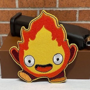 May include: Embroidered patch of a yellow and orange fire spirit with a friendly face. The spirit has large, white eyes and a wide, red smile. The patch is perfect for adding a whimsical touch to clothing, bags, or other accessories.