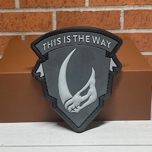 May include: A gray and white embroidered patch with a Mandalorian symbol and the text "This is the Way".