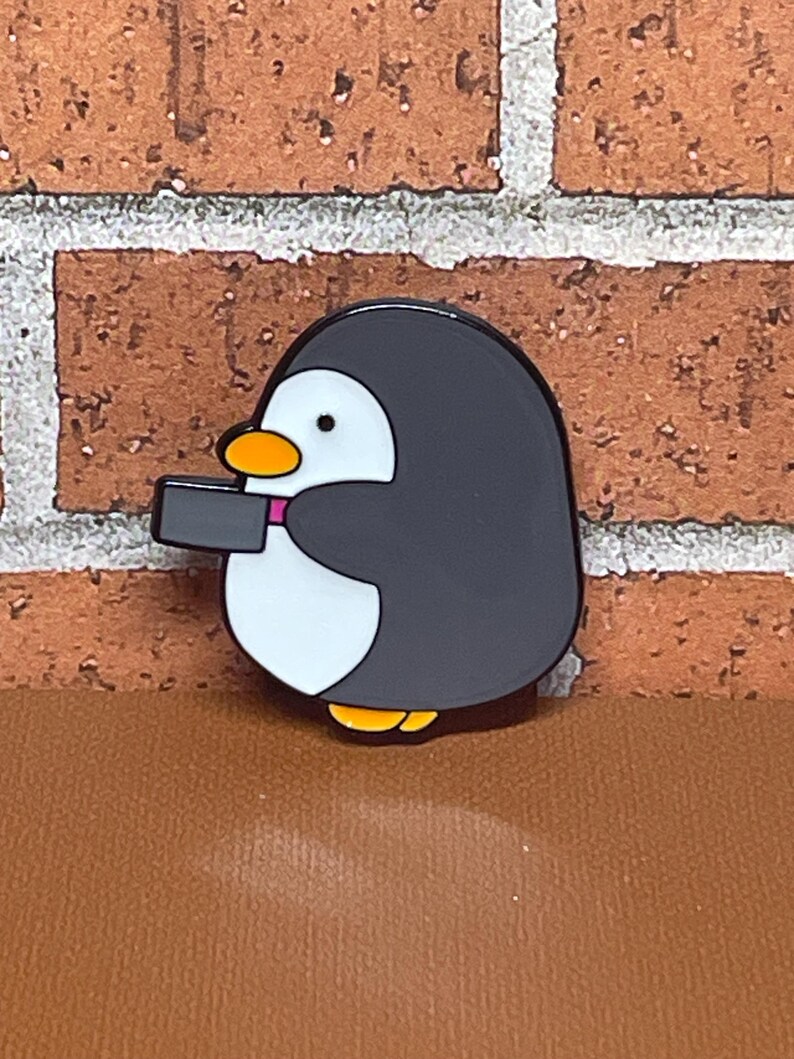May include: A black and white enamel pin of a penguin holding a camera. The penguin has a pink scarf and a yellow beak.