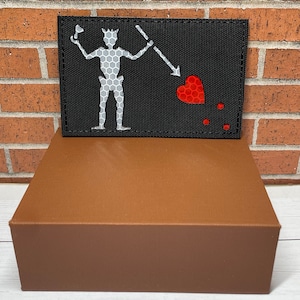 May include: A black patch with a white skeleton figure holding a spear and pointing it at a red heart. There are three red dots below the heart.