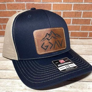 May include: A navy blue and tan trucker hat with a brown leather patch featuring a mountain range and the word "God" in a stylized font.