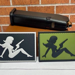 May include: Two black and green patches with a white and black silhouette of a person holding a gun. The patches are on a brown surface.