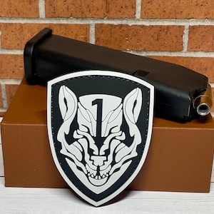 May include: A black and white patch with a wolf's head and the number "1" in the center. The patch is on a brown surface.