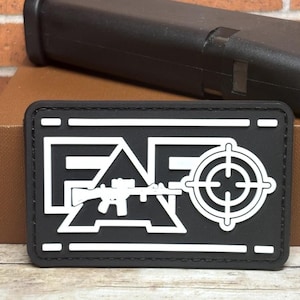 May include: Black rectangular patch with white text and a target reticle design. The patch features the letters "FAG" with a rifle silhouette. A black object is in the background.