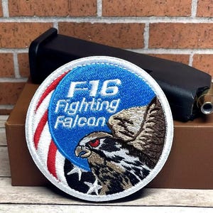 May include: A white, red, and blue embroidered patch with the text "F16 Fighting Falcon" and a falcon illustration.