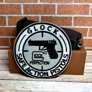 May include: A black and white embroidered patch with the words "Glock Safe Action Pistols" and a silhouette of a handgun. The patch has a gray background with a white circle in the center. The words "Lock Perfection" are printed on the handgun.
