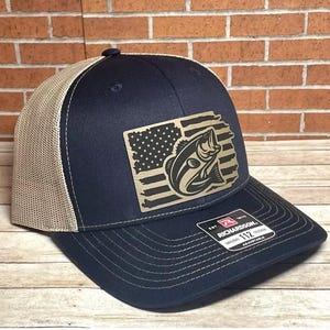 May include: A navy blue and tan trucker hat with a brown patch featuring a bass jumping out of an American flag. The hat is adjustable and has the text "Richardson 112" on the back.