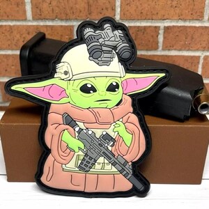 May include: A green and pink character with large ears, wearing a white helmet with a black visor and a pink robe, holding a black rifle. The character is a popular fictional character from the Star Wars franchise.