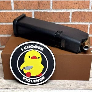 May include: A black and white patch with a yellow cartoon duck holding a knife. The patch has the text "I CHOOSE VIOLENCE" around the duck.
