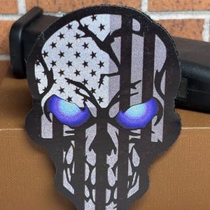 May include: A skull-shaped patch featuring a black and silver American flag design with a cracked texture. The skull has prominent, glowing-effect blue eyes. The design is rendered in a high-contrast style with black outlines and silver details.