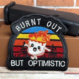 May include: A black embroidered patch featuring a sunset background with orange, yellow, and blue stripes. A cartoon marshmallow with a flame on its head is centered. The text reads BURNT OUT at the top and BUT OPTIMISTIC at the bottom in white.