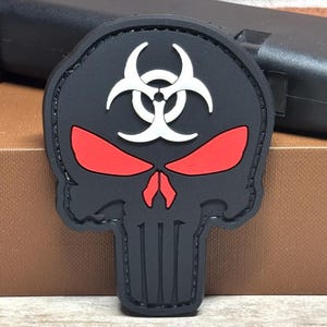 May include: Black skull patch with red eyes and a white biohazard symbol. The patch has a stitched border and a vertical design on the lower part of the skull. The patch is made of a rubbery material.